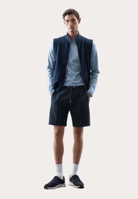 Gilet sportivo blu scuro sopra una maglietta a maniche lunghe azzurra, abbinato a pantaloni corti scuri e sneakers. Calze bianche, design minimalista, senza fantasie.