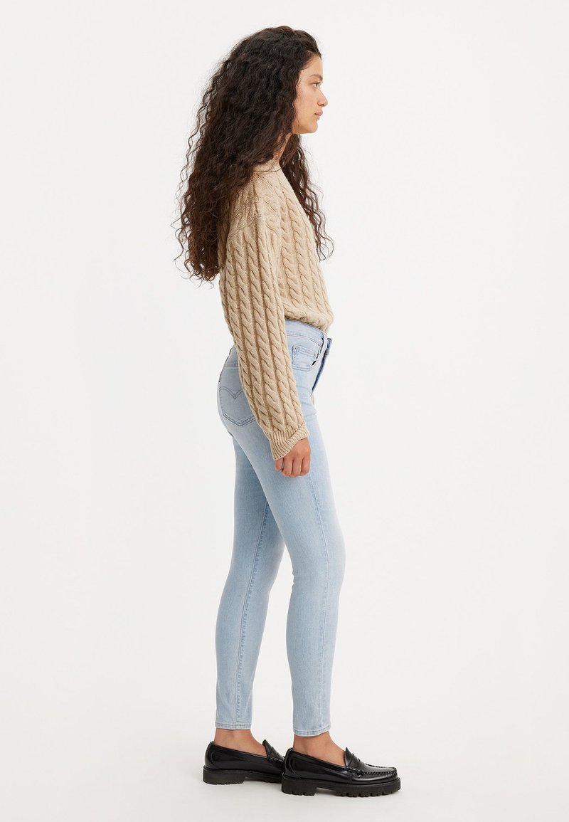 Pull en tricot beige, jean skinny bleu clair et mocassins noirs. La vue latérale met en valeur le tissu texturé et la silhouette ajustée.