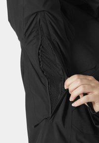 Czarna kurtka z siateczkową podszewką; widoczna ręka otwierająca kieszeń na zamek z boku, przeznaczoną do przechowywania lub wentylacji.