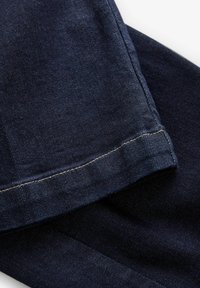 Tejido vaquero azul oscuro doblado para mostrar el detalle del dobladillo cosido y la textura bajo una luz suave sobre un fondo blanco.