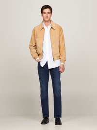 Giacca in suede nocciola con colletto, camicia bianca sotto, jeans blu scuro e scarpe nere. Design semplice, vestibilità casual.