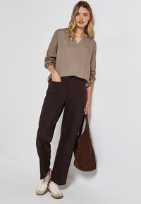 Blouse beige à col en V avec des manches longues, assortie à un pantalon large marron foncé. Le mannequin tient un sac à main en daim marron et porte des baskets blanches.