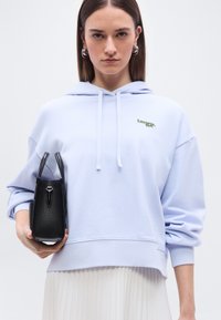 Femme portant un sweat à capuche Lacoste violet clair et une jupe plissée blanche, tenant un sac à main noir texturé sur un fond blanc uni.