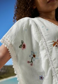 Vit löst sittande topp med blommig broderi i rosa, blått och orange. Spetsdetaljer längs ärmarna ger extra stil och textur.
