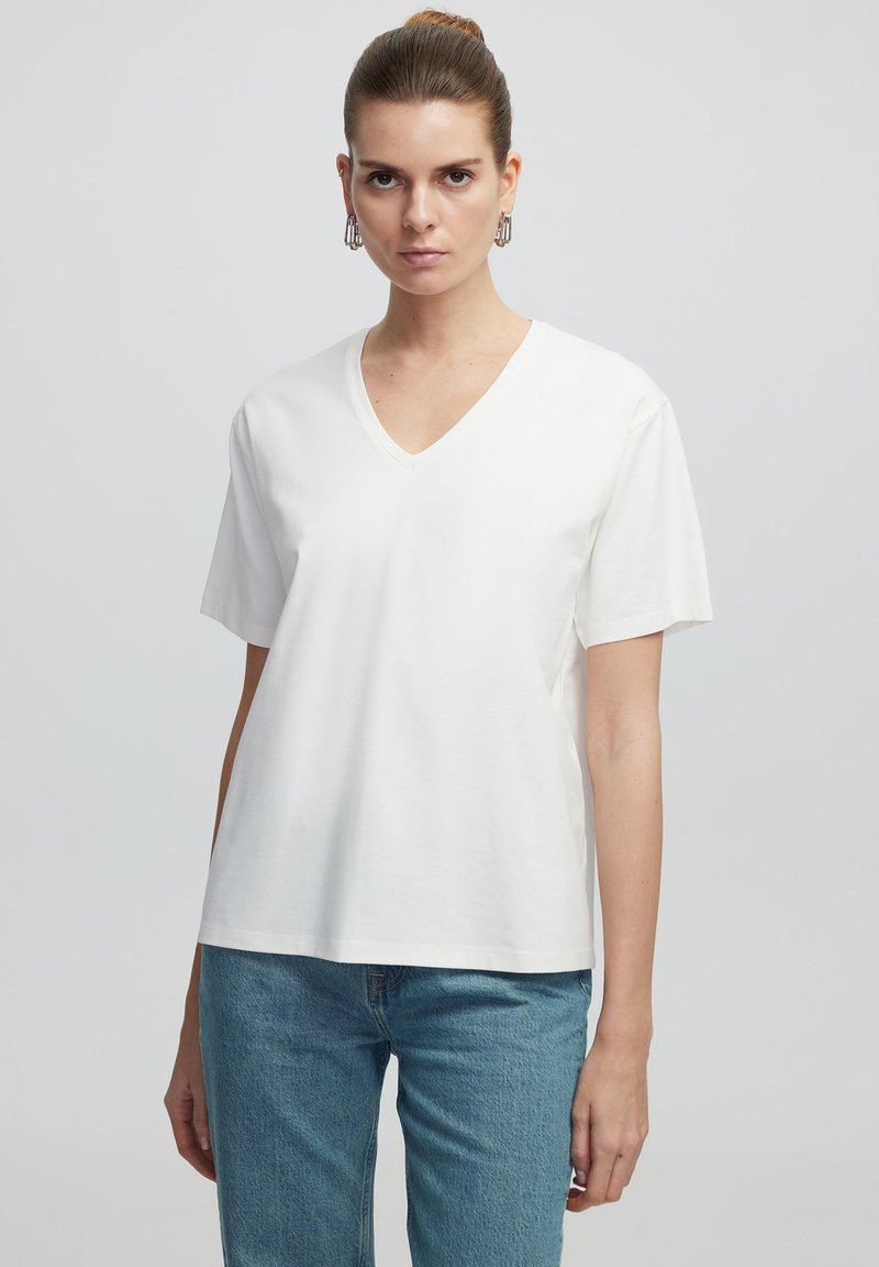 T-shirt bianco in cotone con scollo a V, maniche corte, vestibilità comoda, texture liscia e dettagli di cucitura sottili. Abbinato a jeans azzurri chiari.