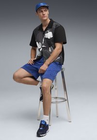 Sort og grå grafisk skjorte med fedt design, blå svedshorts og blå kasket. Iført sorte sneakers med blå accenter og hvide sokker.