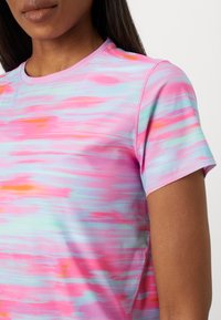 Vrouw draagt een T-shirt met korte mouwen en een ronde hals met een abstract aquarelpatroon in roze, blauw en oranje, zichtbaar van schouders tot borst.