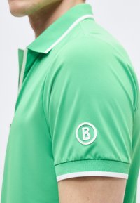 Homme portant un polo vert clair avec un bord blanc et un logo circulaire blanc en forme de "B" sur la manche, sur un fond uni.
