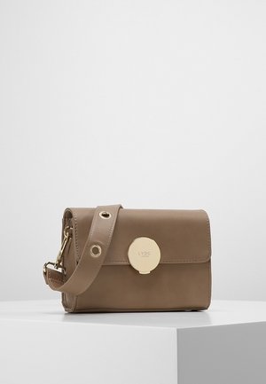 Mała torebka typu crossbody z taupe skóry z szerokim paskiem, złotą okrągłą klamrą i wytłoczonym napisem „LYDC London” na przedniej klapce, umieszczona na białym postumencie.