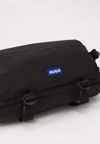 Sort stof rejsetaske med en tekstureret overflade, der har et blåt "HUGO" logo-patch, lynlåslukning og justerbare stropper.