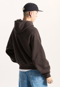 Sudadera con capucha marrón de corte relajado, con dobladillo y puños acanalados. Combinada con jeans de un tono azul claro, que cuentan con bolsillos traseros, y una gorra negra.