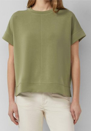 Person wearing an olive green short-sleeve top with centered seam detail and light beige pants against a plain gray background.