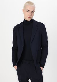 Navy blazer and turtleneck in a smooth fabric, featuring a tailored cut, notched lapels, and side pockets, worn with matching trousers.