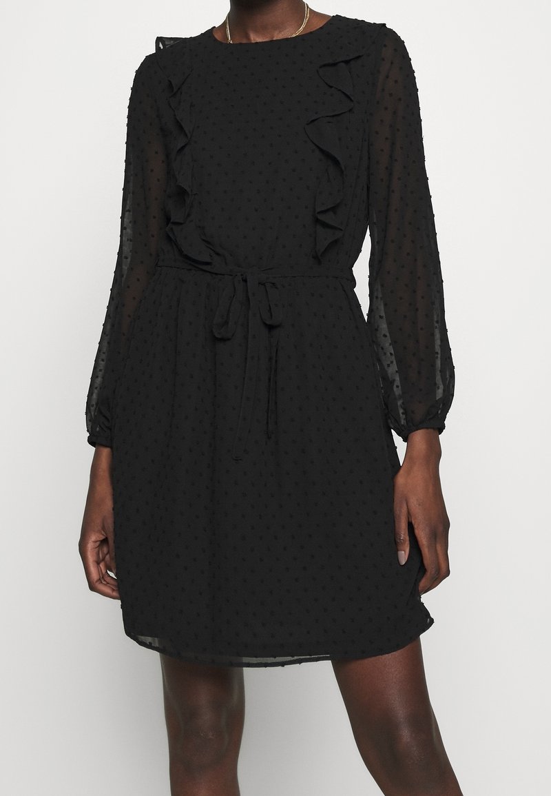 Femme portant une robe noire longueur genoux avec des manches longues transparentes, texture à pois, volants sur la poitrine et une ceinture nouée à la taille.