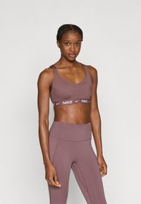 Ανοιχτό καφέ αθλητικό σουτιέν με ντεκολτέ σε σχήμα V και φαρδιές τιράντες, που διαθέτει λωρίδα με το λευκό λογότυπο της Nike. Συνδυάζεται με αντίστοιχα κολάν ψηλόμεσα.