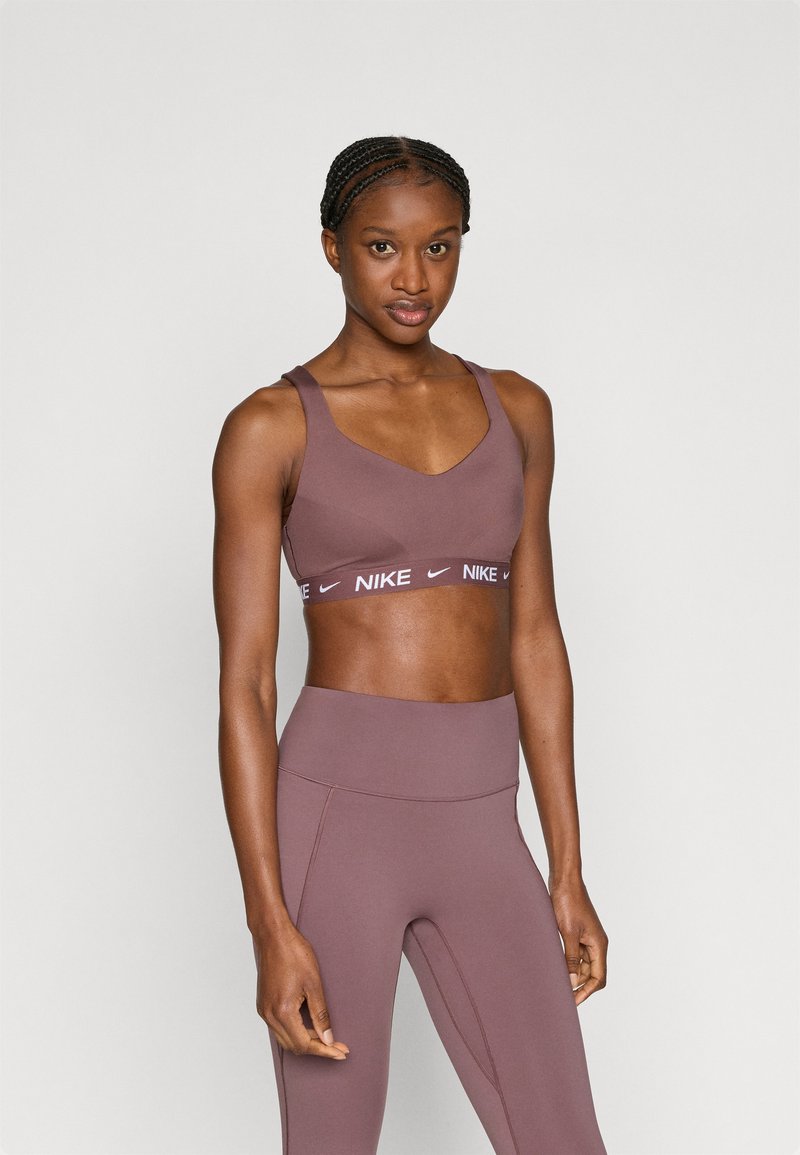 Ανοιχτό καφέ αθλητικό σουτιέν με ντεκολτέ σε σχήμα V και φαρδιές τιράντες, που διαθέτει λωρίδα με το λευκό λογότυπο της Nike. Συνδυάζεται με αντίστοιχα κολάν ψηλόμεσα.