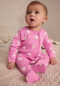 Body côtelé rose avec motif d'étoiles blanches, manches longues et fermeture éclair devant. Texture douce. Bébé assis sur une couverture en tricot de couleur claire.