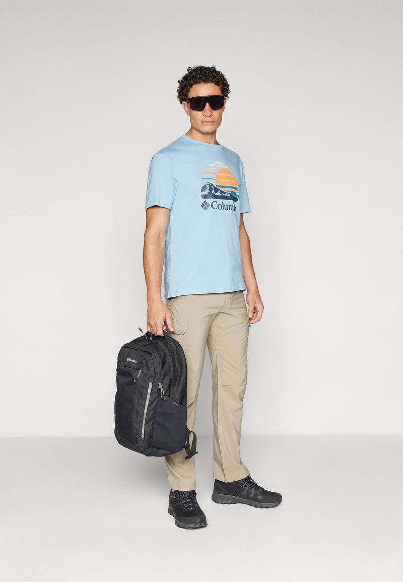 T-shirt bleu clair avec un graphique de coucher de soleil, pantalon kaki, sac à dos noir, lunettes de soleil noires et chaussures de randonnée noires. Un homme se tient debout.
