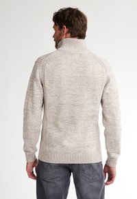 Uomo con capelli castani che indossa un maglione a collo alto beige chiaro in maglia e jeans grigi scuri, in piedi rivolto di spalle su uno sfondo bianco.