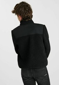 Zwarte fleecejack met een gestructureerde buitenkant, voorzien van een contrasterend glad zwart paneel op de bovenrug en een hoge kraag.