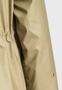 Manche de manteau en tissu beige clair avec détail élastique cousu à la taille et petit logo brodé près du poignet.