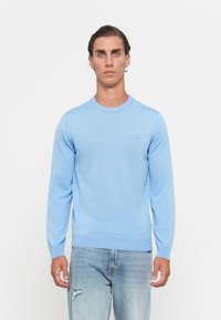 Moški oblečen v svetlo modro dolgosleečno pulover in svetlo modre trganje jeans stojijo ob belem ozadju.