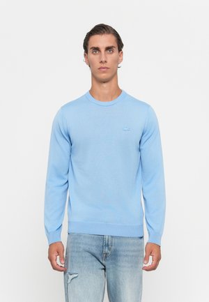 Man in een lichtblauwe lange mouwen trui en lichtblauwe versleten jeans staat tegen een witte achtergrond.