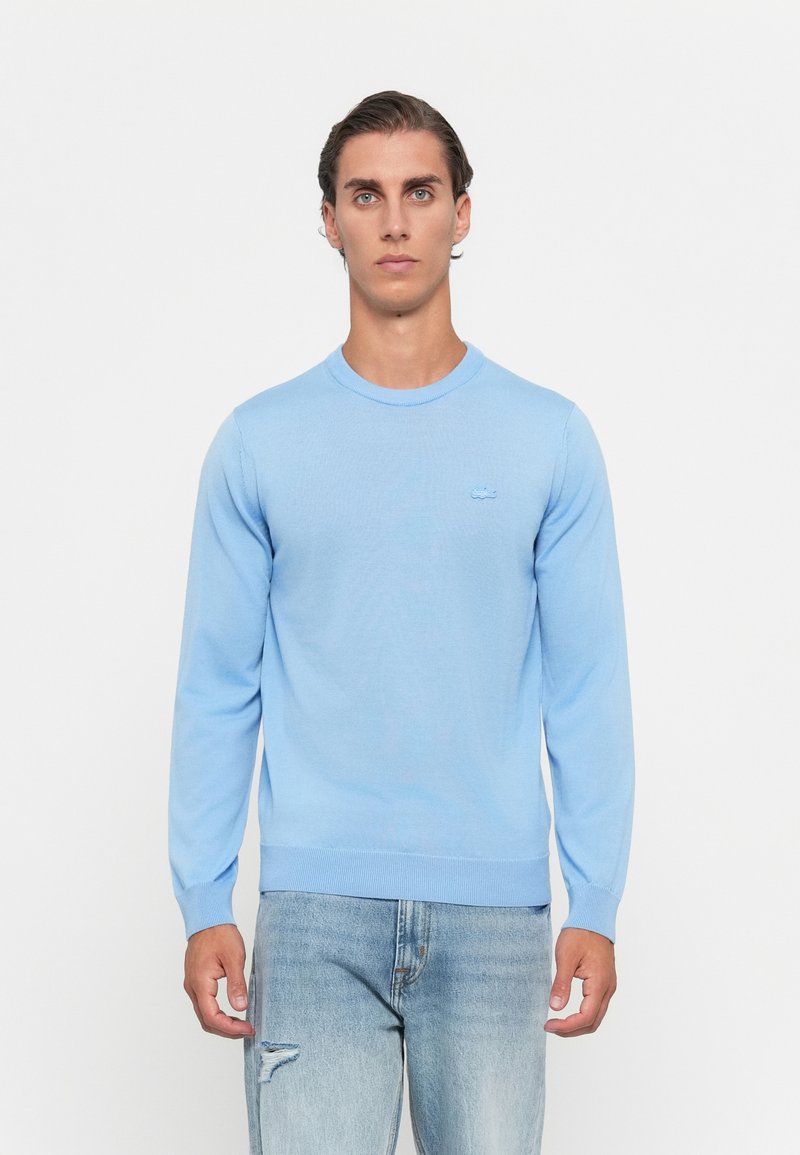 Moški oblečen v svetlo modro dolgosleečno pulover in svetlo modre trganje jeans stojijo ob belem ozadju.