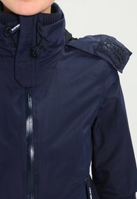 Námořnický větrovka s vysokým límcem, zapínáním na zip a ramenní nášivkou. Má texturovanou tkaninu a boční kapsy se zipy na uzavření.