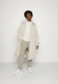 Casaco puffer bege claro com colarinho e bolsos laterais, usado sobre uma camisa branca e calças de treino bege. Ténis brancos completam o visual.