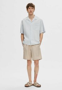 Koszula z krótkim rękawem w jasnoniebieską kratę z kieszonką, zestawiona z beżowymi szortami i brązowymi sandałami na płaskiej podeszwie z paskowym wykończeniem.