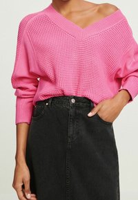 Rosa gestrickter Pullover mit V-Ausschnitt und überschnittenen Schultern, kombiniert mit einem schwarzen Denim-Rock mit hoher Taille. Strukturiertes Material und lockere Passform.