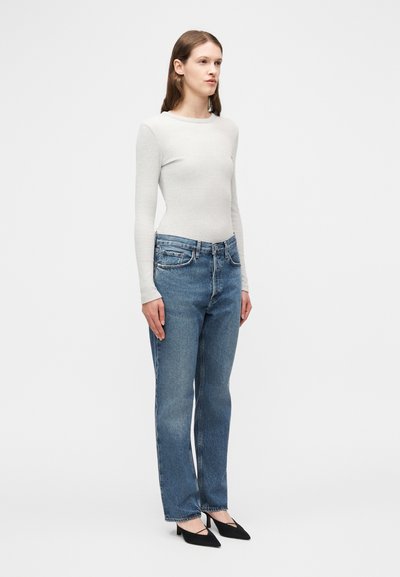 Γυναίκα που στέκεται φορώντας εφαρμοστή ανοιχτογκρι μακρυμάνικη μπλούζα, τζιν παντελόνι με ίσια γραμμή και μαύρα μυτερά παπούτσια με χαμηλό τακούνι, μπροστά σε λευκό φόντο.