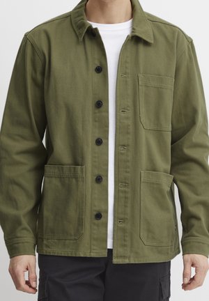 Mand iført olivengrøn jakke med knapper og tre forlommer over en hvid skjorte og sorte bukser, stående mod hvid baggrund.