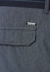 Dunkel gemusterter Stoff mit einem karrierten Design. Verfügt über eine quadratische Tasche und ein Metalletikett, das "CABA MODA" am Bund trägt.