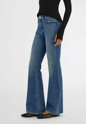 Donna che indossa jeans blu a vita alta a zampa, maglia nera a maniche lunghe e ballerine nere con punta, in piedi davanti a uno sfondo semplice.