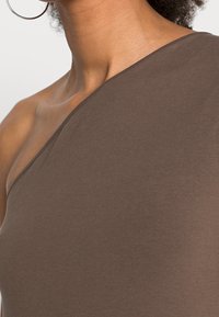 Hnědý top na jedno rameno vyrobený z hladkého, elastického materiálu; má přiléhavý design a minimalní akcenty. Detailní pohled odhaluje strukturu a střih.