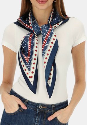 Kobieta w białym dopasowanym t-shircie i niebieskiej wzorzystej chustce zawiązanej wokół szyi, z rękami w kieszeniach niebieskich dżinsów.