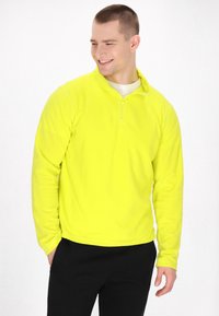 Giacca a pullover con zip di un brillante giallo, realizzata in un tessuto morbido e leggero. Presenta maniche lunghe e un colletto alto. Indossata con pantaloni neri.