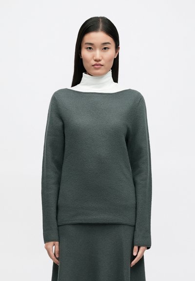 Σκούρο πράσινο πουλόβερ από υφασμάτινη ύφανση, με φαρδιά λαιμόκοψη και μακριά μανίκια, φορεμένο πάνω από λευκό ζιβάγκο. Απλός, μοντέρνος σχεδιασμός.