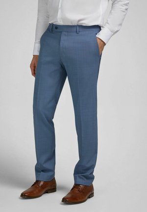 Homme portant un pantalon de costume bleu clair, une chemise blanche et des chaussures habillées en cuir marron, debout avec une main dans la poche.