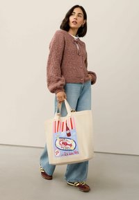 Pletený kardigan v svetloružovej farbe s textúrovaným vzorom a uzáverom na mašľu; béžová kabelka s potlačou homára, pruhovanými akcentmi a pevným uchom.