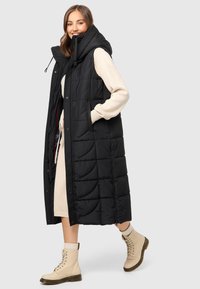 Zwarte, gewatteerde lange vest met een capuchon, voorzien van zijzakken en een rits over de volle lengte. Draag het over een gebreide crèmekleurige jurk en beige veterschoenen.