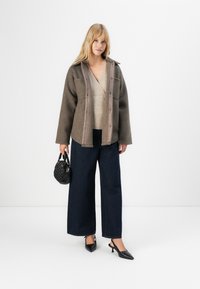 Vrouw in een bruine getextureerde jas over een beige V-hals trui, donkere wijd uitgesneden jeans, zwarte puntige hakken, met een kleine zwarte handtas.
