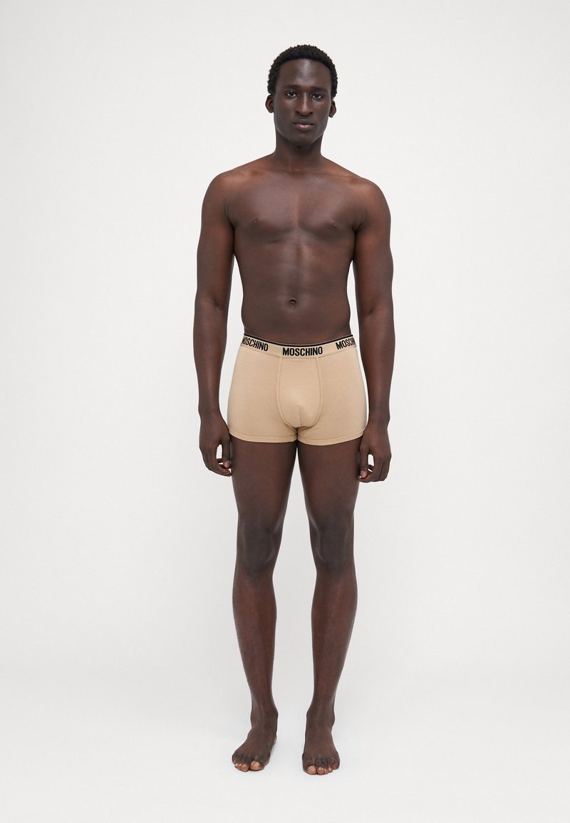 Beige bokszernadrág fekete Moschino logós derékkal, puha anyagból készült, egy személy viseli, aki egyenes háttér előtt áll.