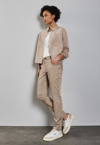 Chaqueta de ante beige con dos bolsillos en el pecho, camiseta blanca, pantalones ajustados beige claros y zapatillas blancas con detalles en color marrón.