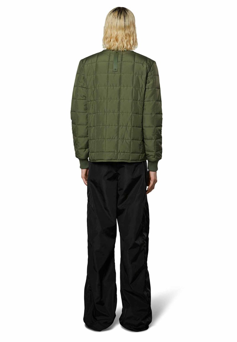 Giacca trapuntata verde oliva con polsini a coste, indossata sopra pantaloni neri larghi, che mostrano una superficie testurizzata e un design minimalista.