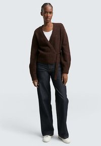 Bruine overslagtrui met een gestructureerde afwerking en een ceintuur in de taille. Gecombineerd met donkere wijd uitlopende jeans en witte sneakers.