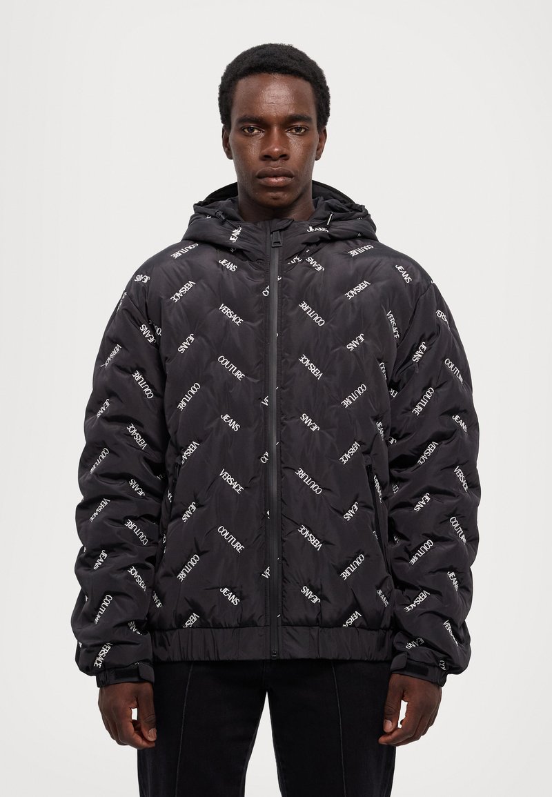 Chaqueta acolchada negra con textura acolchada, que cuenta con una capucha y marcas en blanco en contraste en toda la prenda. Cierre de cremallera con puños acanalados.