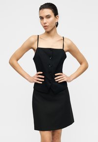 Μαύρο αμάνικο φόρεμα με εφαρμοστή μπλούζα, τετράγωνη λαιμόκοψη και πέντε κουμπιά. Η ευθεία φούστα φτάνει μέχρι το μέσο του μηρού. Ομαλή υφή.